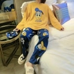 Schattige Disney Donald damespyjama in mosterdgeel en blauw gedragen door een vrouw zittend op een bed in een huis