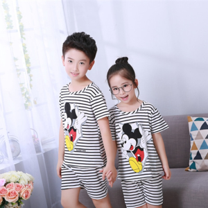 Zwart-wit gestreepte zomerpyjama met Mickey Mousse opdruk voor kinderen gedragen door een kleine jongen en een klein meisje voor een stoel in een huis
