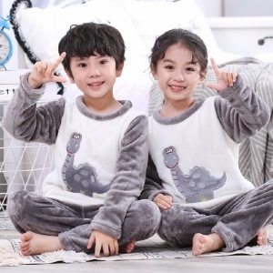 Tweedelige grijze flanellen pyjama met dinosaurusmotief voor kinderen gedragen door een kleine jongen en een klein meisje in een huis