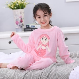 Flanellen pyjama met lange mouwen en zeemeerminpatroon voor een meisje, gedragen door een klein meisje op een bed in een huis