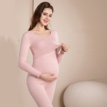 Zwangere brunette vrouw die een roze zwangerschapspyjama draagt, ze raakt met één hand haar ronde buik aan en trekt met de andere aan de bovenkant van haar pyjama